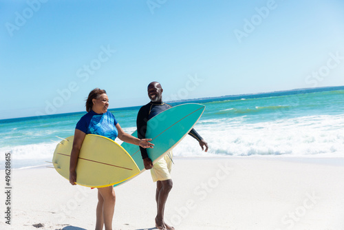 African american senior couple carrying surfboards walking at beach against blue sky with copy space