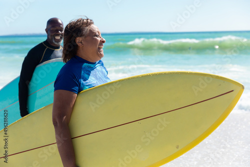 Cheerful african american senior couple carrying surfboards walking at beach on sunny day