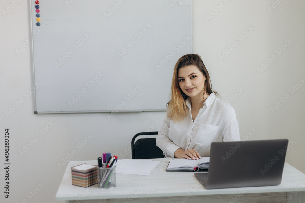 Portrait of a beautiful young teacher conducting online learning in the classroom for her students. The girl is dressed in a white shirt. She looks at the camera Portrait of a beautiful young teacher conducting online learning in the classroom for her students. The girl is dressed in a white shirt. She looks at the camera