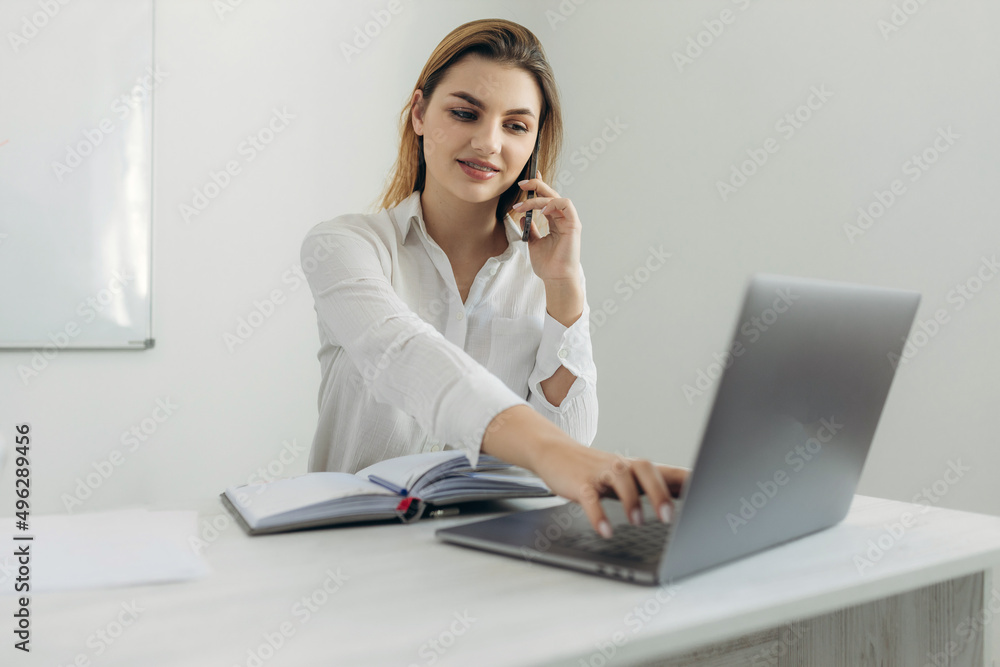 Portrait of young business woman talking on smartphone working at laptop and writing in notebook at home office. Beautiful girl at the computer table at home. Portrait of young business woman talking on smartphone working at laptop and writing in notebook at home office. Beautiful girl at the computer table at home.