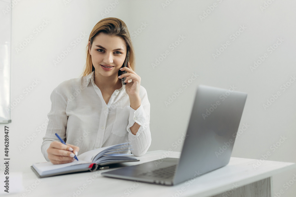Portrait of young business woman talking on smartphone working at laptop and writing in notebook at home office. Beautiful girl at the computer table at home. Portrait of young business woman talking on smartphone working at laptop and writing in notebook at home office. Beautiful girl at the computer table at home.
