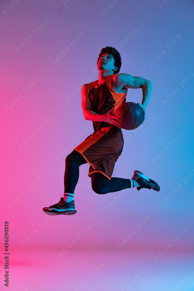 Sportive man playing basketball isolated on gradient pink blue studio background in neon light. Youth, hobby, motion, activity, sport concepts.