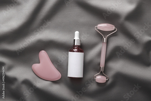 Serum bottle, jade massage roller with guasha stone on gray silk background