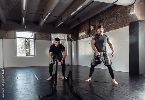 Functional training. Two male athletes are training with a kettlebell and battle ropes in a sports hall