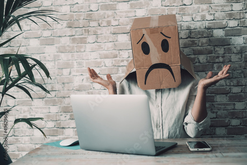 Modern worker with carton sad box on head open arms with desolation gesture in front of a laptop. Online crypto smart working business job activity. Concept of fail and lost work. Bad day office photo