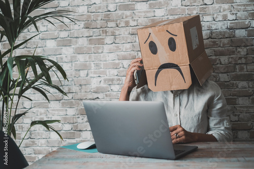 People at work with sad carton box on head diong phone call in front of a laptop on the desk. Sad worker anonymous. Business fail activity and unhappy leisure activity online. Idendity lost photo