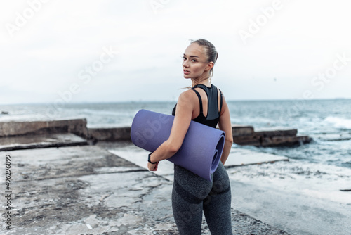 Fototapeta Naklejka Na Ścianę i Meble -  Portrait of young fit woman in sportswear with yoga mat on urban beach