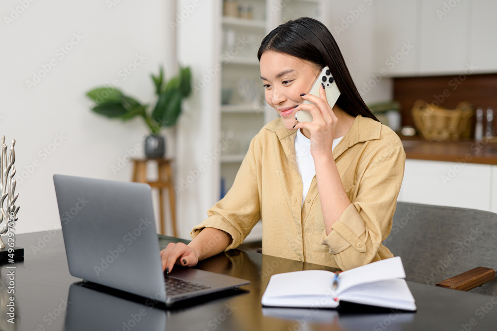Girl works remotely. Multitask asian freelance woman talks on the smartphone and takes notes with pen, using laptop for remote work from home, write down some information sitting at the table Girl works remotely. Multitask asian freelance woman talks on the smartphone and takes notes with pen, using laptop for remote work from home, write down some information sitting at the table