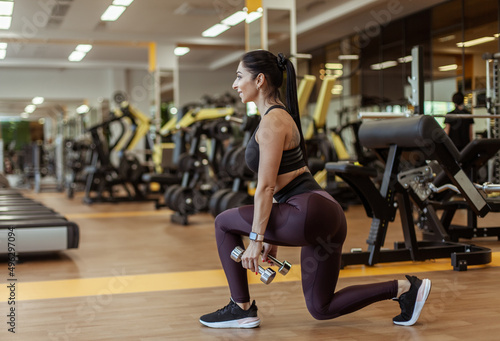 Athletic fit woman in sportswear trains leg muscles doing lunges with dumbbells in her hands in the gym