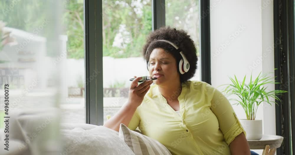 Video of happy plus size african american woman with headphones sitting on sofa with smartphone Video of happy plus size african american woman with headphones sitting on sofa with smartphone