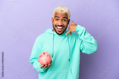 Young Colombian man holding a piggybank isolated on purple background making phone gesture. Call me back sign