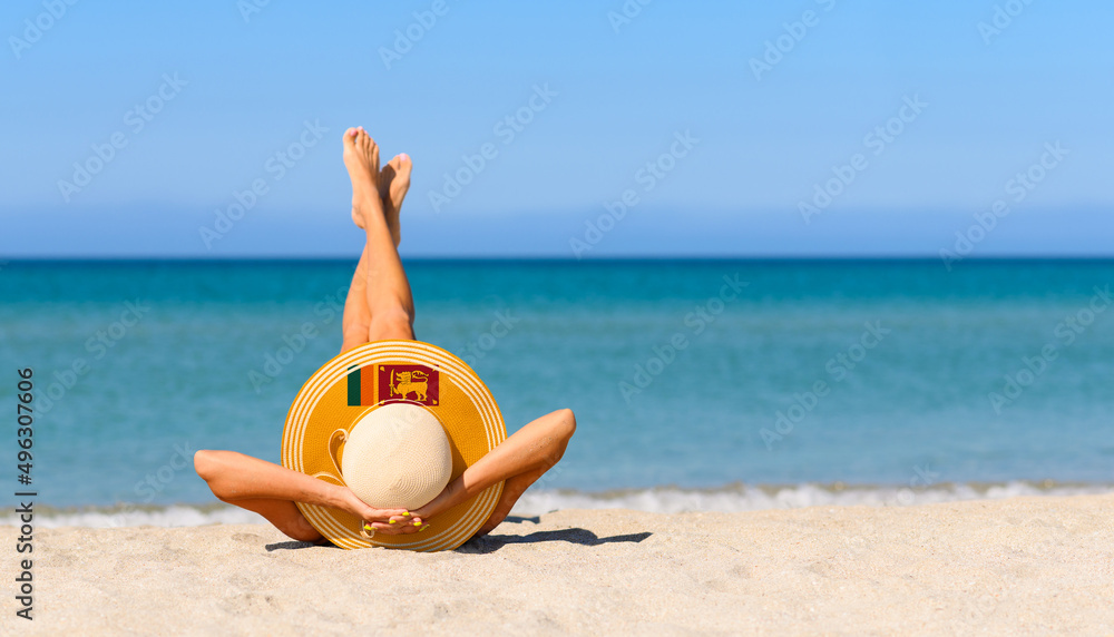 Fototapeta premium A slender tanned girl on the beach in a straw hat in the colors of the flag of Sri Lanka. The concept of a perfect vacation in a resort in the Sri Lanka. Focus on the hat.