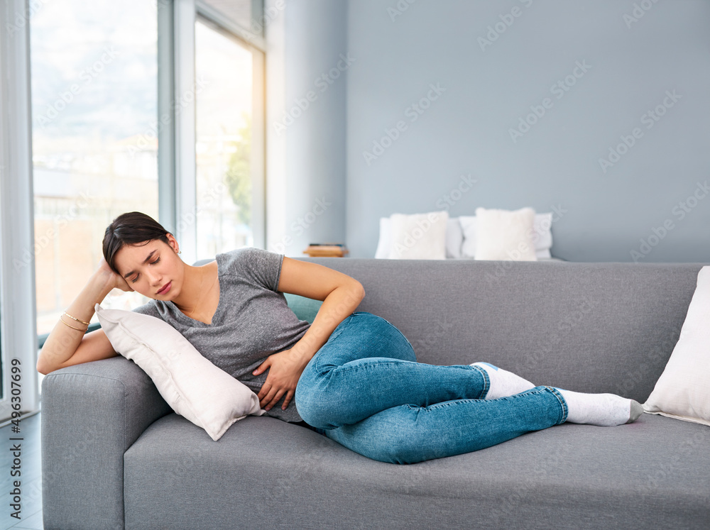 I think Im going to take easy today. Shot of a uncomfortable looking young woman holding a water bottle tightly while trying to get rest on a sofa at home. I think Im going to take easy today. Shot of a uncomfortable looking young woman holding a water bottle tightly while trying to get rest on a sofa at home.