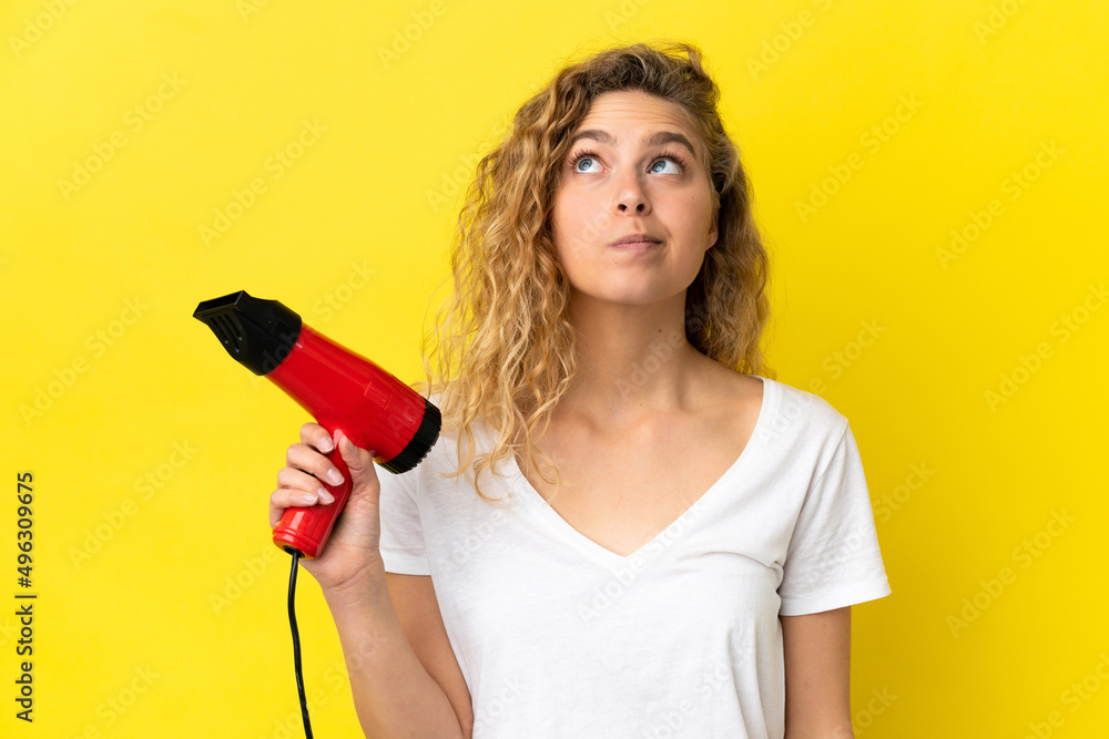 Young blonde woman holding a hairdryer isolated on yellow background and looking up Young blonde woman holding a hairdryer isolated on yellow background and looking up