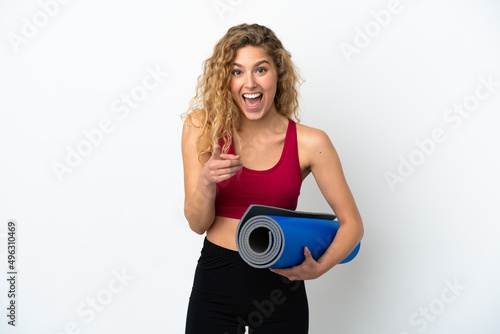 Young sport blonde woman going to yoga classes while holding a mat isolated on white background surprised and pointing front
