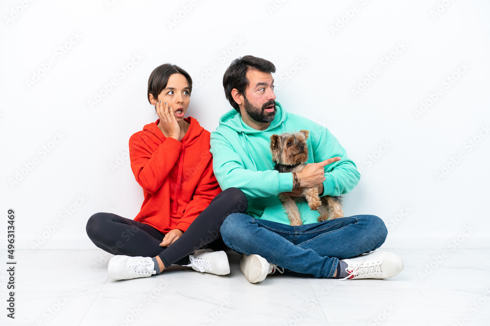 Young caucasian couple sitting on the floor with their pet isolated on white background pointing finger to the side with a surprised face Young caucasian couple sitting on the floor with their pet isolated on white background pointing finger to the side with a surprised face