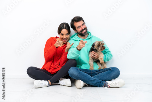 Young caucasian couple sitting on the floor with their pet isolated on white background points finger at you with a confident expression