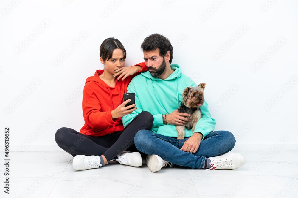 Young caucasian couple sitting on the floor with their pet isolated on white background reading a message with the mobile Young caucasian couple sitting on the floor with their pet isolated on white background reading a message with the mobile