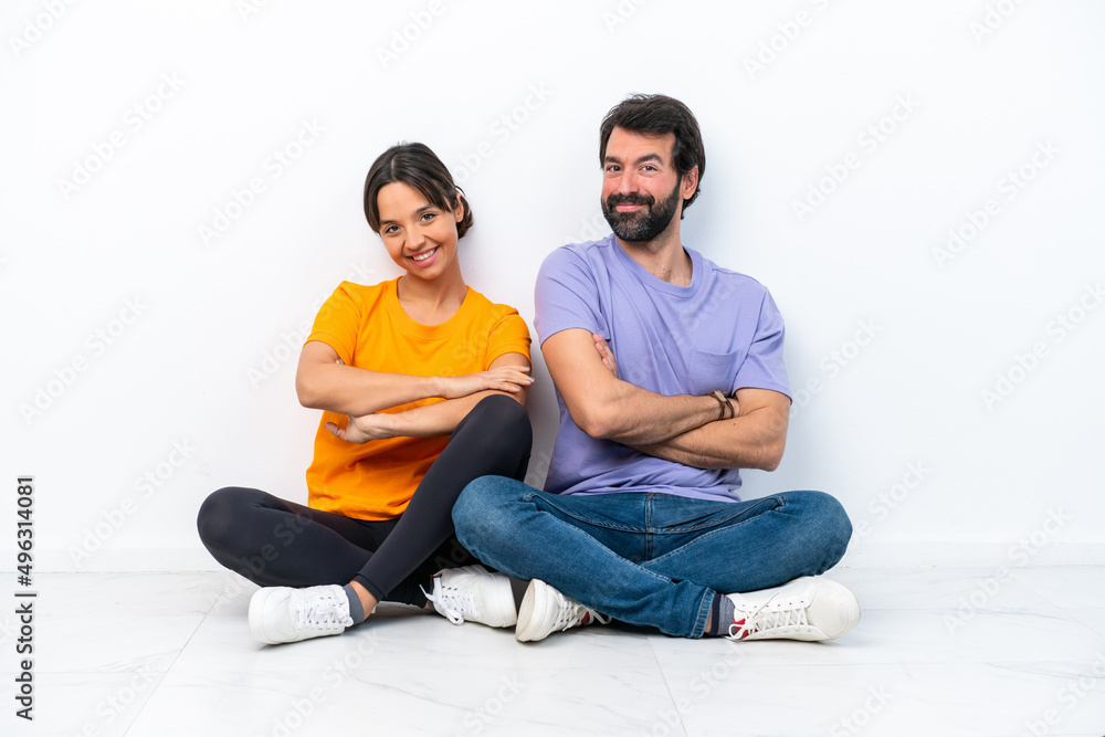 Young caucasian couple sitting on the floor isolated on white background keeping the arms crossed in lateral position while smiling Young caucasian couple sitting on the floor isolated on white background keeping the arms crossed in lateral position while smiling