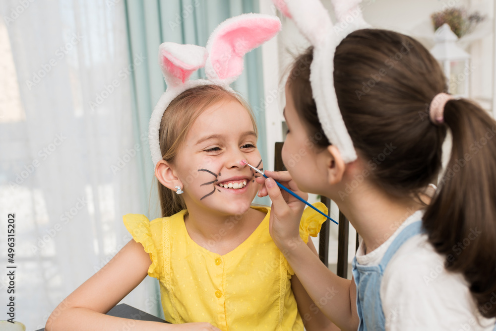 two girls celebrate Easter, paint eggs with a brush. Happy girls smile and laugh while drawing on their face. A cute little girl in bunny ears is getting ready for the holiday. two girls celebrate Easter, paint eggs with a brush. Happy girls smile and laugh while drawing on their face. A cute little girl in bunny ears is getting ready for the holiday.