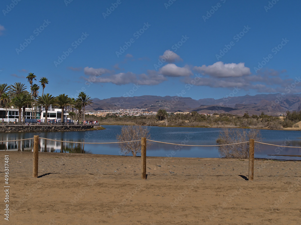 View of small lagoon La Charca in Meloneras, part of Maspalomas, southern Gran Canaria, Spain with fence in front and tourists walking on promenade.