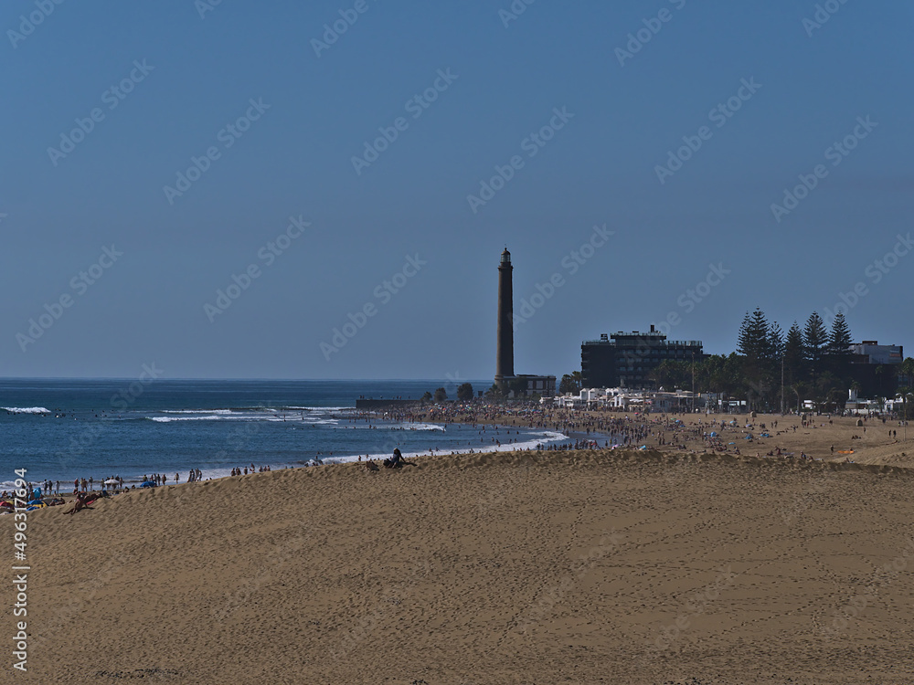 Fototapeta premium Aerial view over crowded beach Playa de Maspalomas with sand dune in front and lighthouse Faro de Maspalomas in the south of Gran Canaria, Spain.