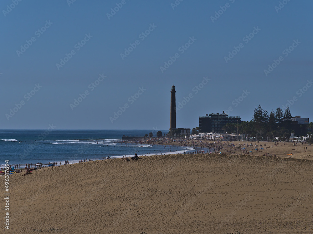 Aerial view over crowded beach Playa de Maspalomas with sand dune in front and lighthouse Faro de Maspalomas in the south of Gran Canaria, Spain.