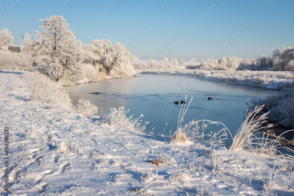 Winter scenery in a village. Frosty morning in a village near the lake. Snow cover on the land and trees. Small mist over water of the lake.