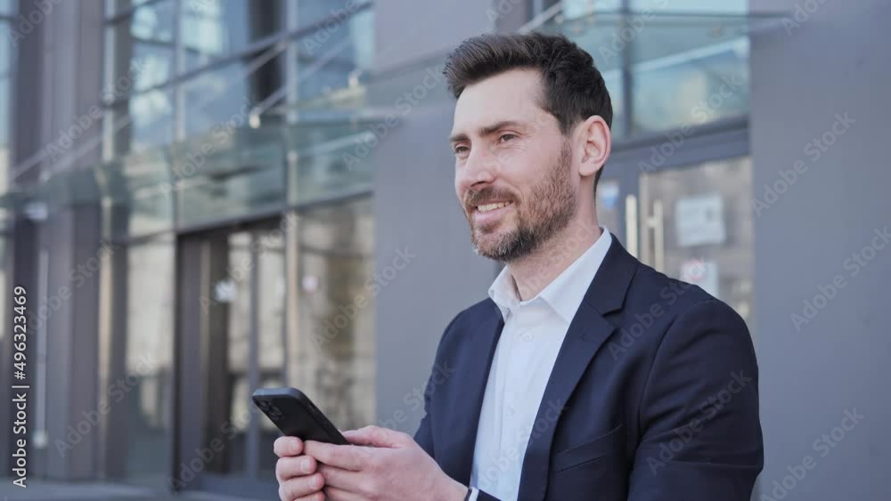 Handsome Businessman sitting on the bench near big modern Business district. Young bearded man texting Messages on Smartphone Man scrolling typing taping browsing data internet social media smiling. Handsome Businessman sitting on the bench near big modern Business district. Young bearded man texting Messages on Smartphone Man scrolling typing taping browsing data internet social media smiling.