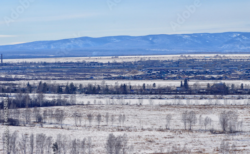 The landscape of the river valley, along which a small town is located. The blue mountains of Sayana are visible on the horizon. Cloudy day. Nature of Eastern Siberia in March