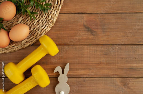 Fototapeta Naklejka Na Ścianę i Meble -  Two heavy dumbbells, decorative Easter bunny, eggs and boxwood branches. Healthy fitness lifestyle composition, gym workout and training concept. Fit flat lay with copy space on wooden background.