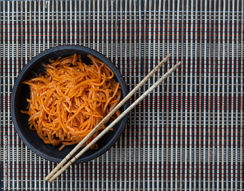 Korean carrots in a brown clay bowl on a dark bamboo background. Chopsticks on a bowl. Top view, space for text