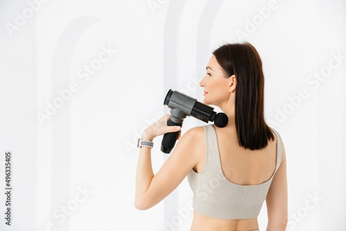 A girl in a beige sports suit massaging her back. vibrating massager close-up. Electric massager pistol in hand massaging the muscles. Sports recovery concept. 