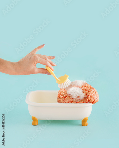 Fotografie Minimal abstract concept made of  young female hand brushing model of human brain in the tub with foam, on isolated pastel aquamarine blue background