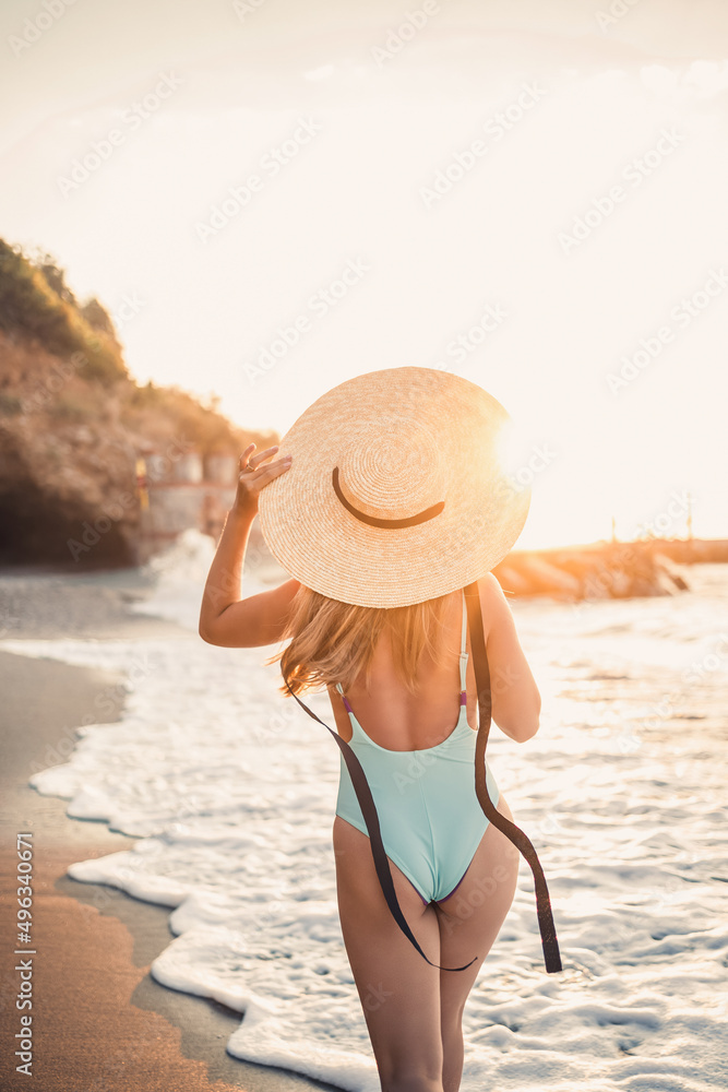 A young tanned woman in a beautiful swimsuit with a straw hat stands and rests on a tropical beach with sand and looks at the sunset and the sea. Selective focus. Vacation concept by the sea