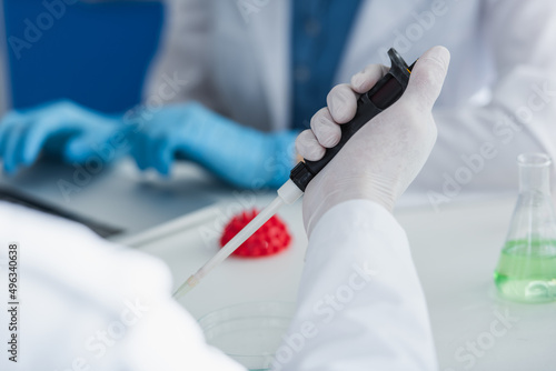 cropped view of scientist in latex glove holding micropipette near blurred model of coronavirus bacteria.