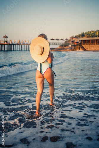 A woman in a swimsuit and a hat with glasses walks along the beach at sunset. The concept of sea recreation. Selective focus