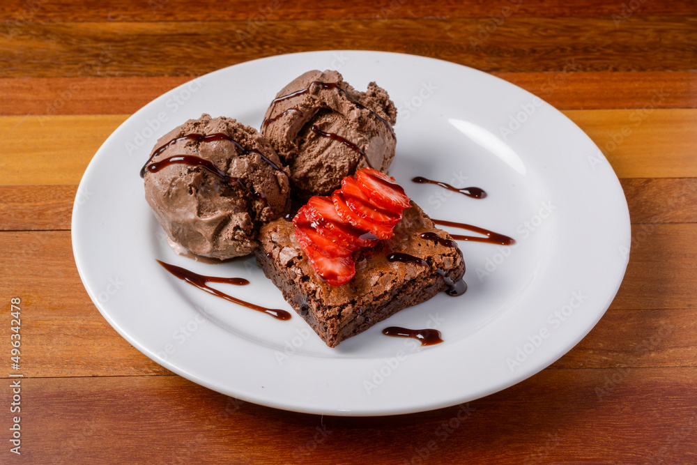 Brownie with sliced strawberries and chocolate ice cream on a white plate on a wooden table.