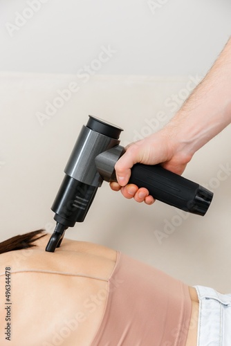 A therapist massages a woman's back with a massage percussion device in her home. The therapist's hand holds a therapeutic vibrating massager. Physical therapy and muscle recovery and massage