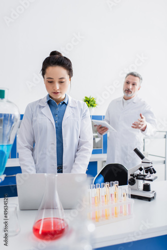 blurred bioengineer talking to colleague using laptop near microscope and test tubes.