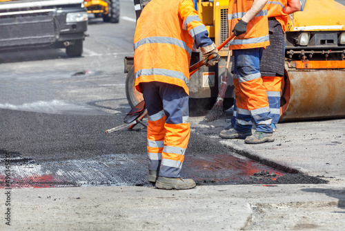 A group of road workers in orange overalls are repairing a section of the carriageway by rolling up fresh asphalt.