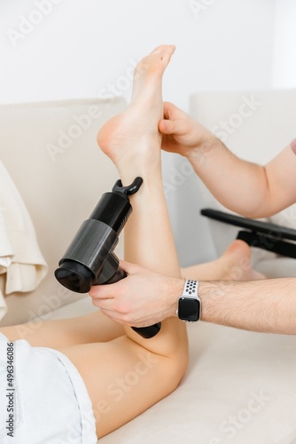 A therapist massages a woman s leg with a massage percussion device in her home. The therapist s hand holds a therapeutic vibrating massager. Physical therapy and muscle recovery and massage