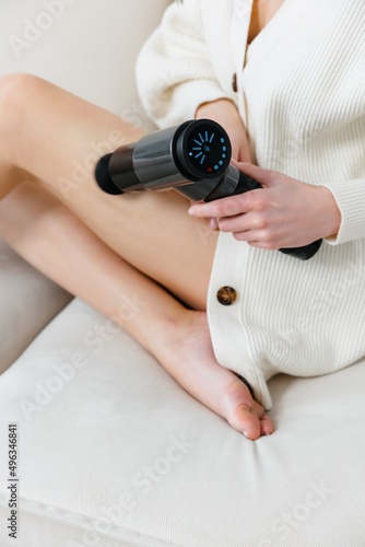 A girl at home holds a therapeutic percussive vibrating massage gun in her hand and massages her muscles.  Sports recovery concept after a workout. 