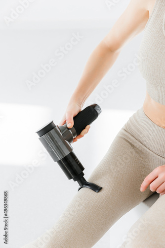 A girl at home on the couch massaging with a vibrating massager. An electric therapeutic pistol massager in her hand massages her leg muscles. Sports recovery concept after a workout. 