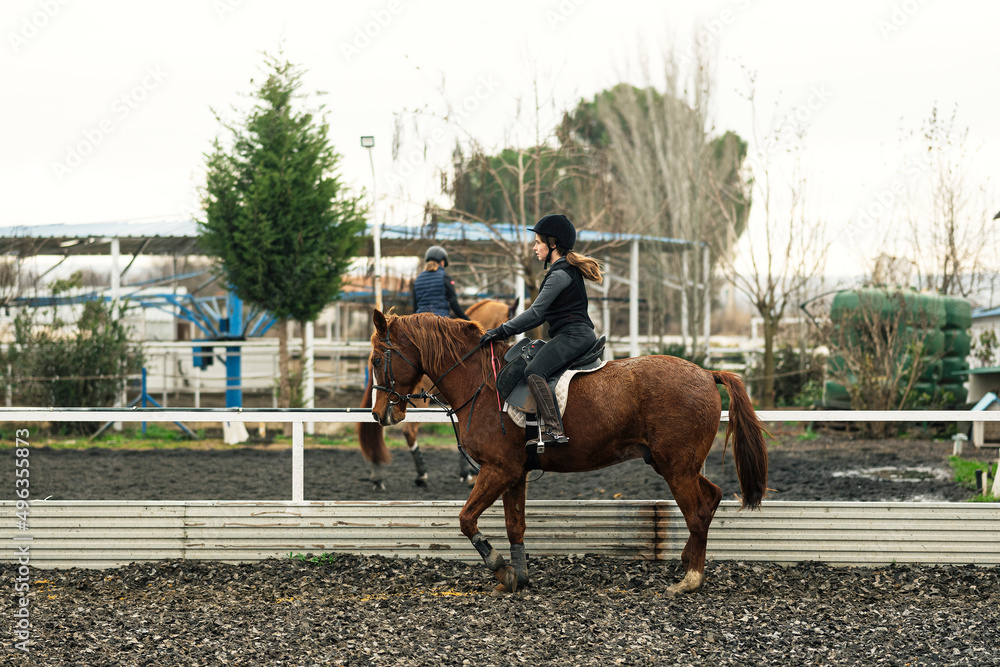 Young girl in helmet and special clothes riding a horse on the sandy ground of the paddock Young girl in helmet and special clothes riding a horse on the sandy ground of the paddock