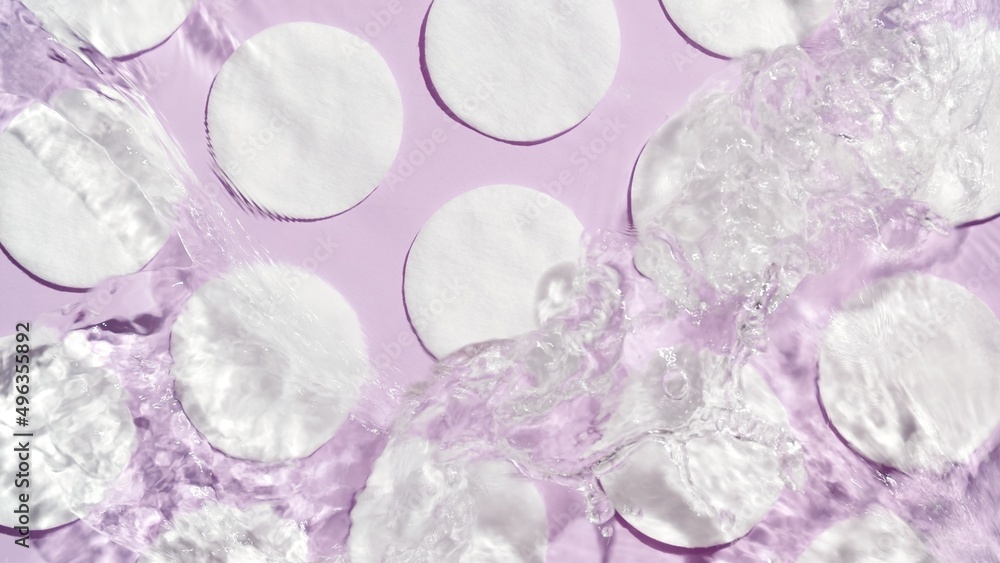 Poured water splashing and making ripples over cotton pads arranged in rows on purple background | product background, cleansing lotion commercial
