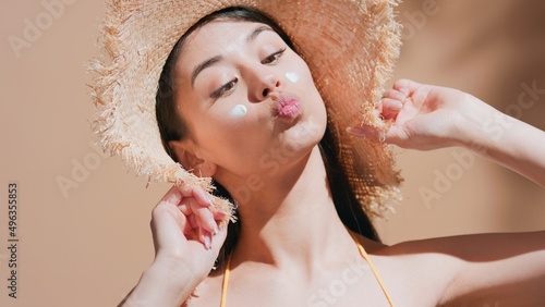 Close-up beauty portrait of young dark-haired asian woman with sunscreen on her cheeks in a straw hat enjoys the sun and grimacing | Skin care concept