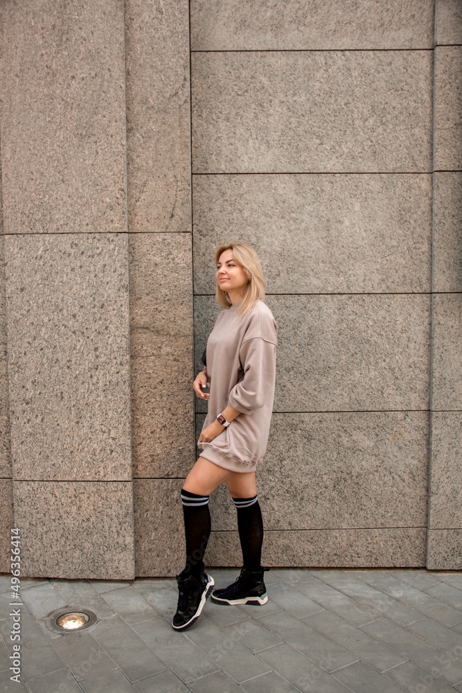 sports blonde girl in beige oversize dress is standing casual in profile with smile near street beige store building background and looking away with hand near hair. lifestyle concept, free space
