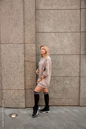 sports blonde girl in beige oversize dress is standing casual in profile with smile near street beige store building background and looking away with hand near hair. lifestyle concept, free space