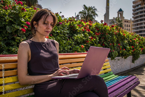 Successful Thirtysomething Woman Working Remotely on her Pink Laptop from a Park Bench Painted with Rainbow Colors of the LGBT Flag photo